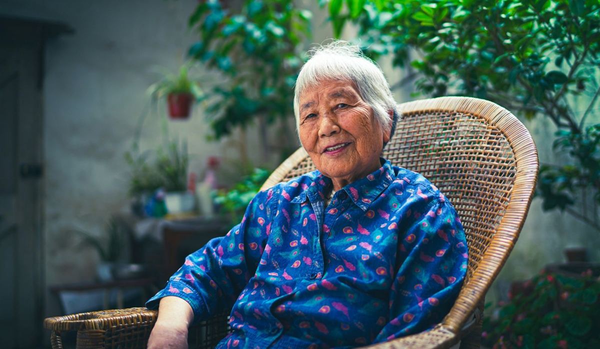 An older woman smiling in a wicker chair surrounded by plants in her courtyard.
